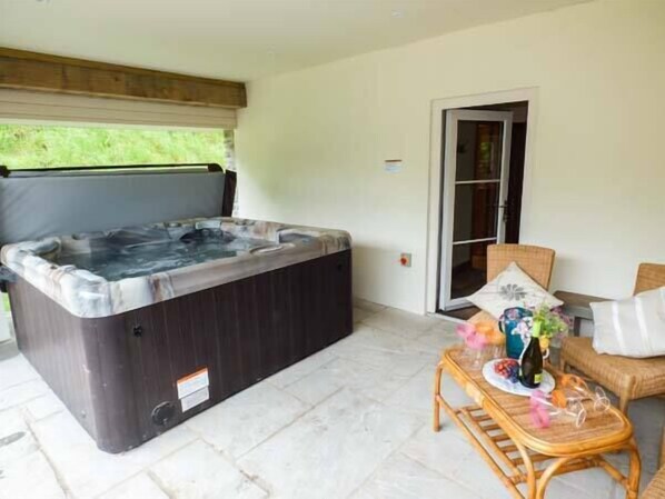 A hot tub in a covered outdoor area with a wicker chair and a small table with bottles and glasses. - The Old Mill (Llandrindod Wells)