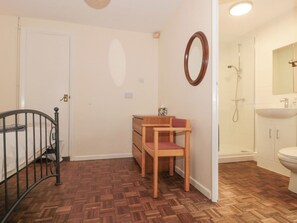 A room with a wooden floor, a bed with a metal frame, a wooden chair, a small wooden table with a tissue box, a mirror on the wall, and a bathroom with a sink and shower. - The Cottage (Watchet)