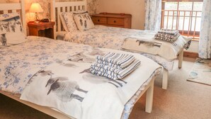 A bedroom with two beds, floral wallpaper, a wooden dresser, a nightstand with a lamp, and a window with curtains. - Stable Barn (Ashbourne)