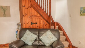 A brown leather sofa with two decorative pillows, a wooden staircase, and a wall-mounted shelf with books and a vase. - Stable Barn (Ashbourne)