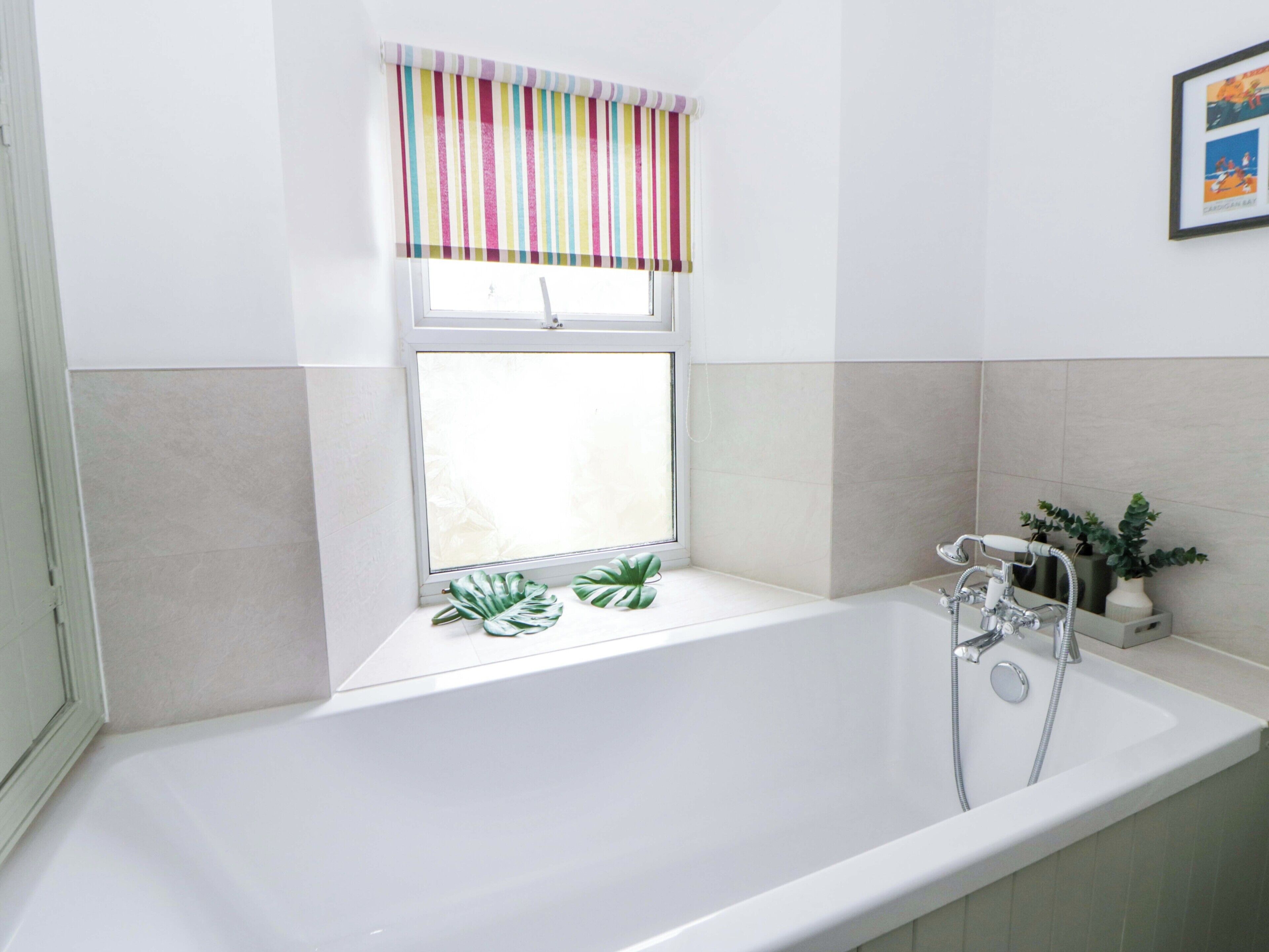 Ein Badezimmer mit einer weißen Badewanne, einem Fenster mit gestreiftem Rollo und einem Blumentopf auf der Fensterbank.