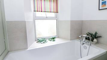Ein Badezimmer mit einer weißen Badewanne, einem Fenster mit gestreiftem Rollo und einem Blumentopf auf der Fensterbank.