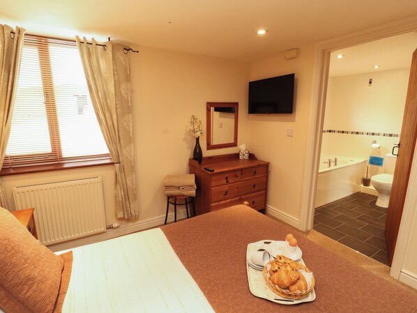 Habitación de hotel con cama, tocador con espejo, televisor en la pared y baño con bañera e inodoro.