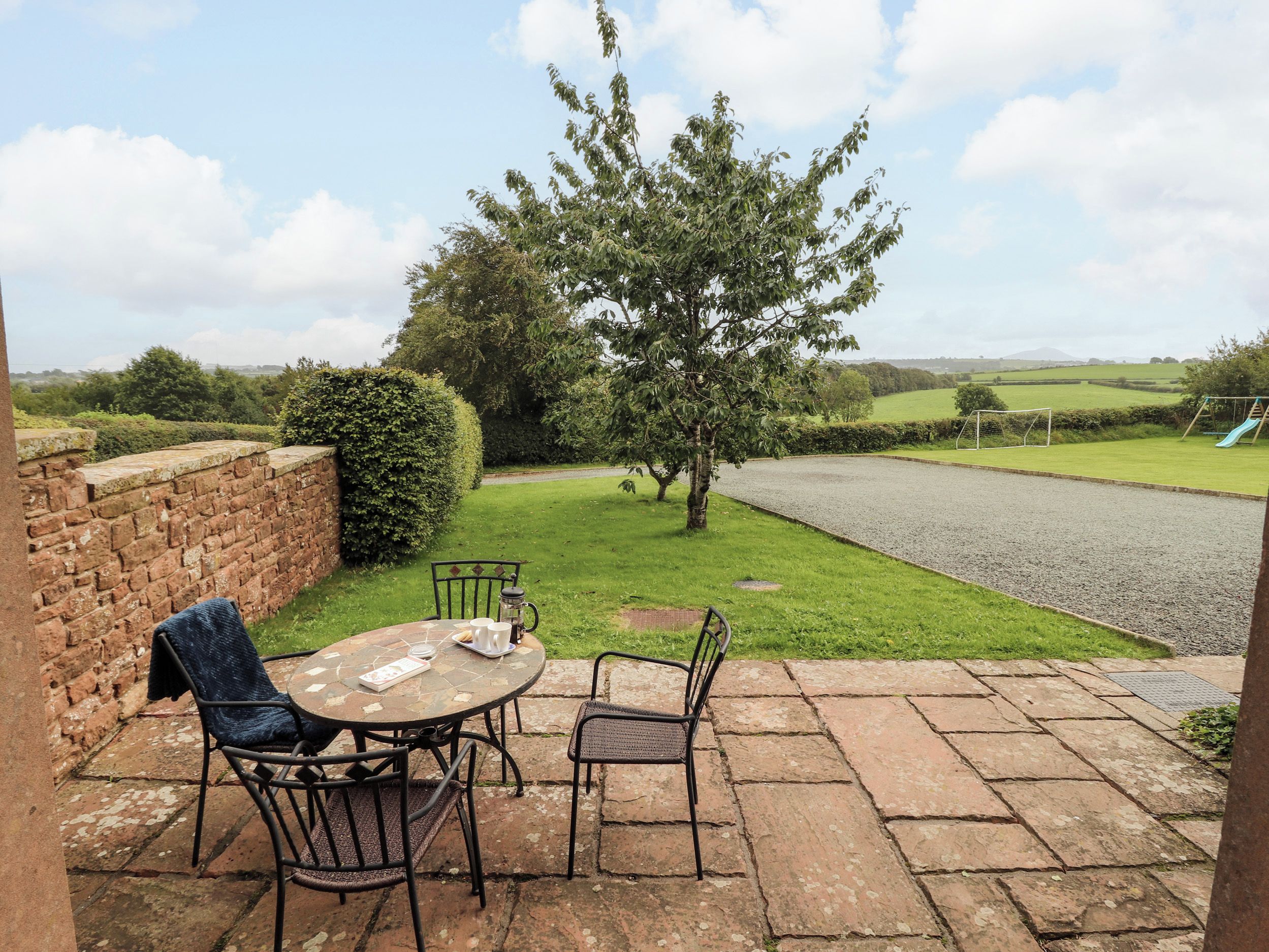 An outdoor patio with a table and chairs, a brick wall, and a view of a grassy field and a playground.