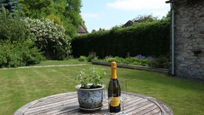 A round wooden table with a bottle of Prosecco and two glasses, a potted plant, and a stone wall in a garden setting. - The Studio (Ashbourne)