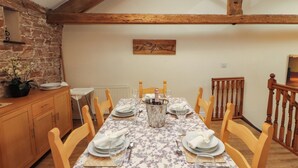 A dining area with a long table set for four, wooden chairs, a stone wall, and a wooden ceiling beam. - Hayloft Cottage (Wigton)