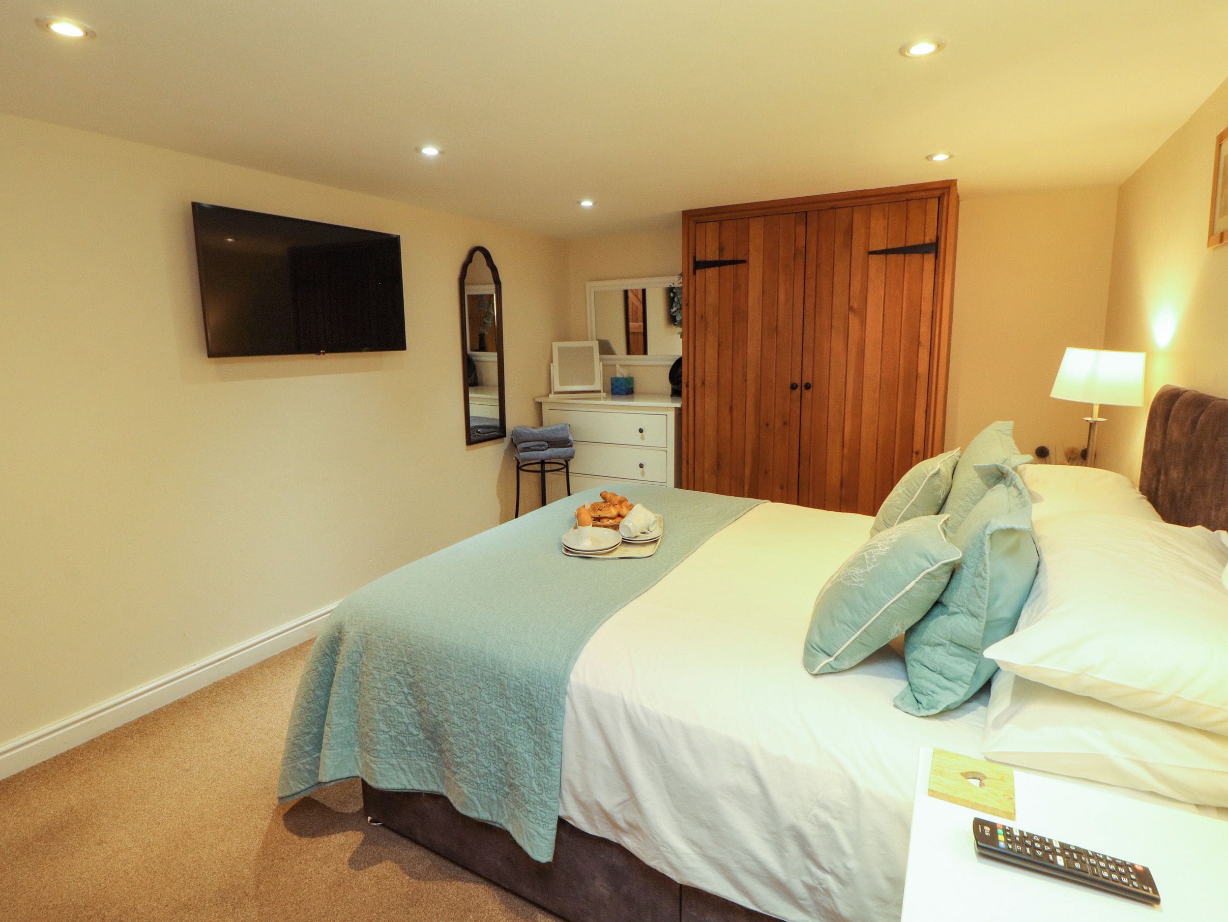 A hotel room with a bed, a television mounted on the wall, a desk with a chair, a wardrobe, and a bedside table with a lamp.