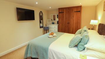 A hotel room with a bed, a television mounted on the wall, a desk with a chair, a wardrobe, and a bedside table with a lamp.