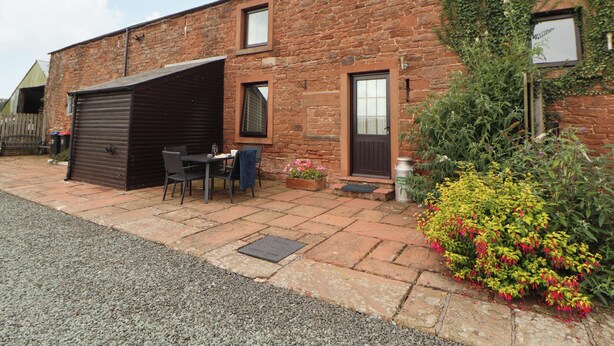 A brick building with a garage, a small patio with a table and chairs, and a garden with flowers. - Hayloft Cottage (Wigton)