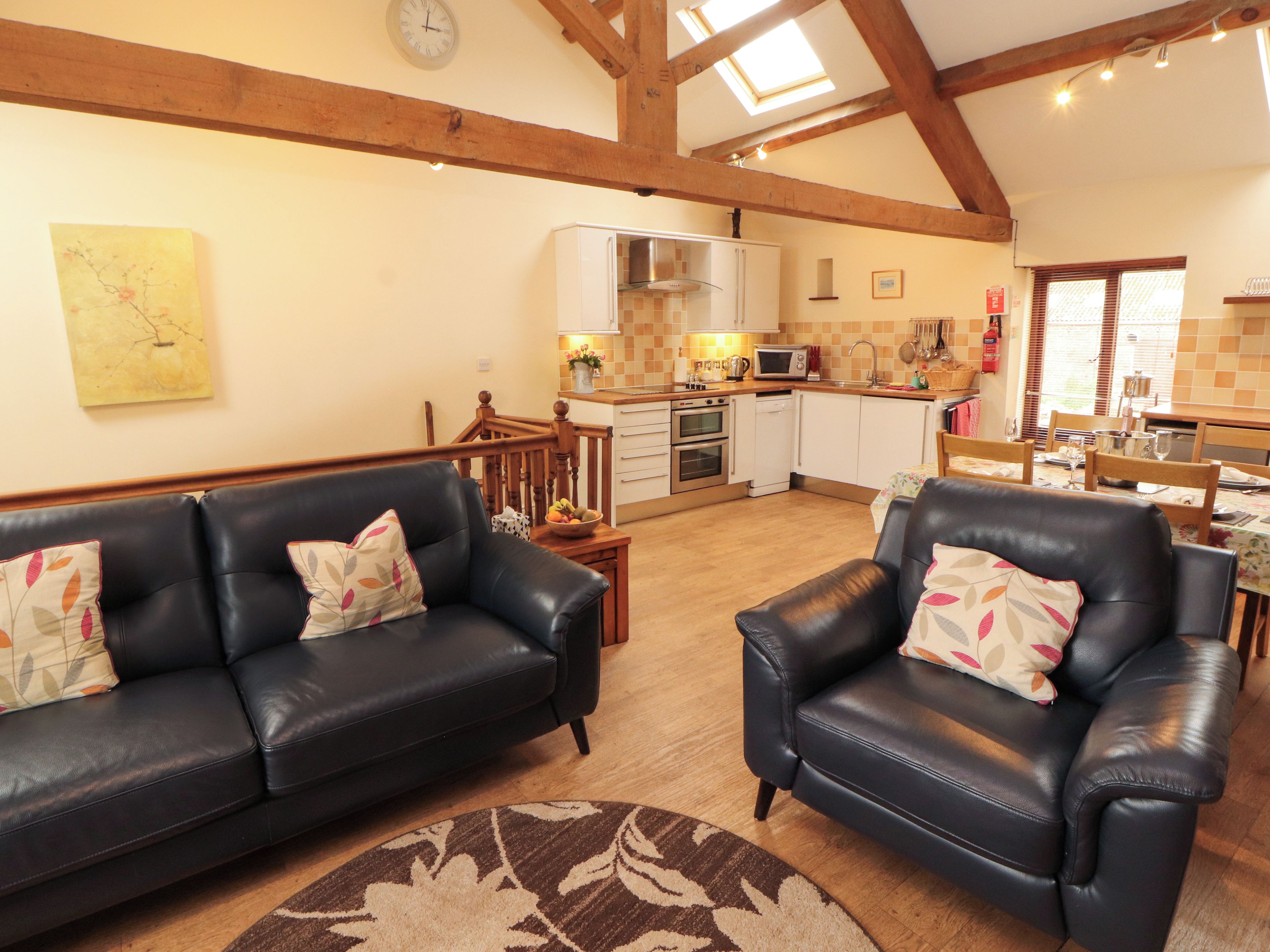 A living room with a black leather sofa and armchair, a wooden ceiling, and a kitchen area with white cabinets and a microwave.
