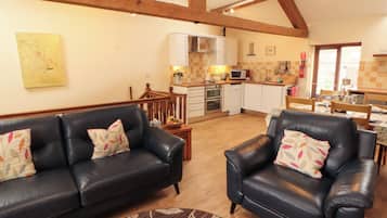 A living room with a black leather sofa and armchair, a wooden ceiling, and a kitchen area with white cabinets and a microwave.