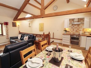 A cozy living room with a black leather sofa, wooden dining table set for two, and a kitchen area with white cabinets and a tiled backsplash. - Horseshoe Cottage (Wigton)