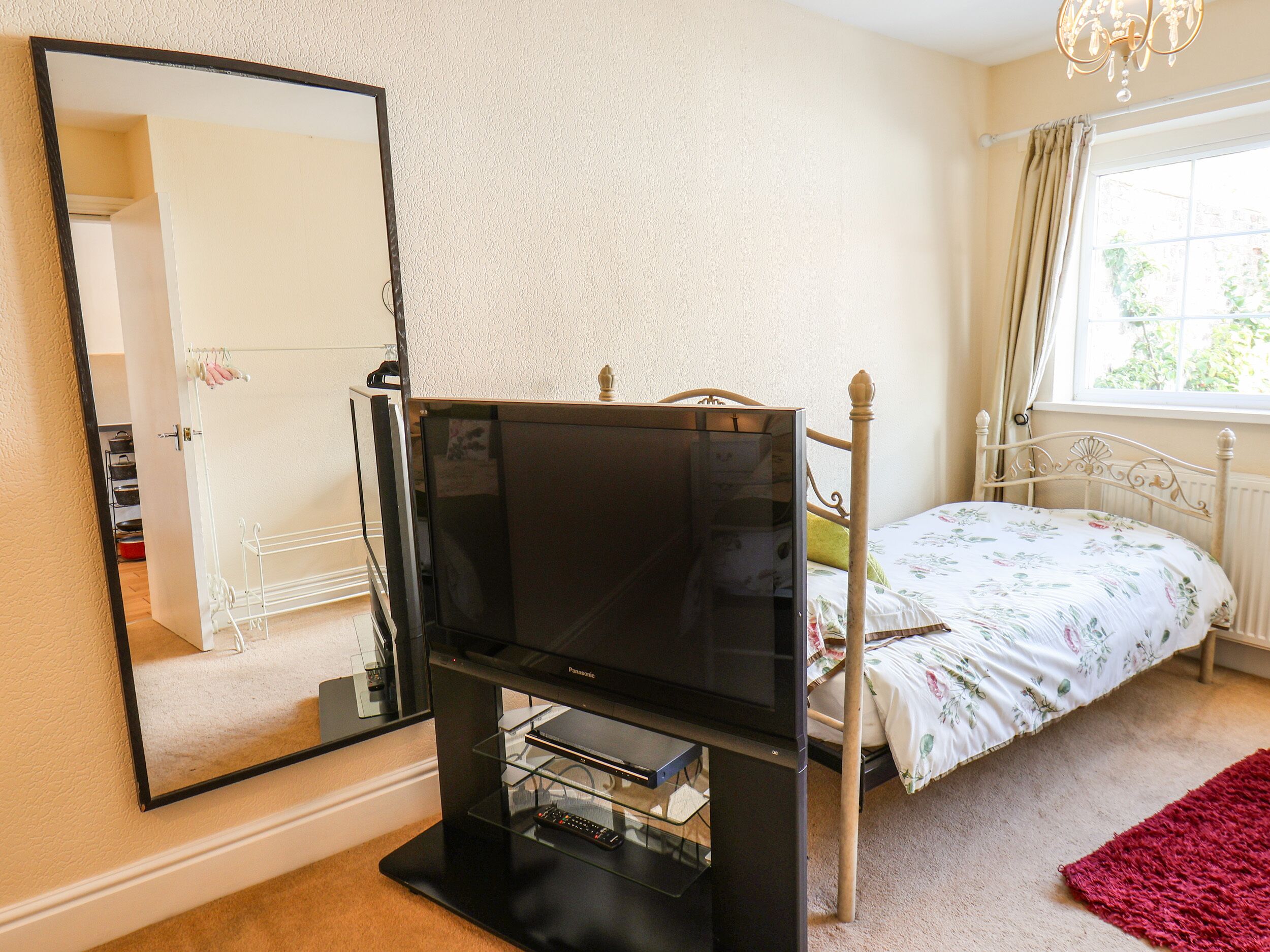 Een slaapkamer met een bed, een televisie op een standaard, een spiegel en een raam met gordijnen.