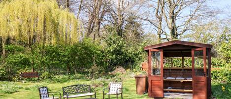 A gazebo with a bench and chairs in a garden with trees and a bench in the foreground.