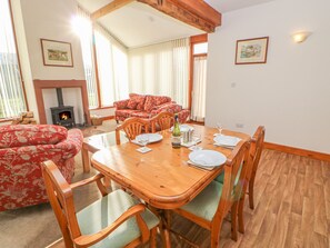 A dining area with a wooden table set for four, surrounded by wooden chairs with green cushions. A fireplace is visible in the background, and there are two framed pictures on the walls. - Swift Cottage (Brampton)