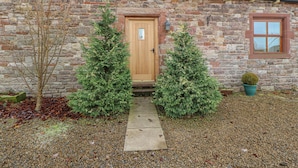 A stone building with a wooden door and a window. There are two evergreen trees in front of the door. - Swift Cottage (Brampton)