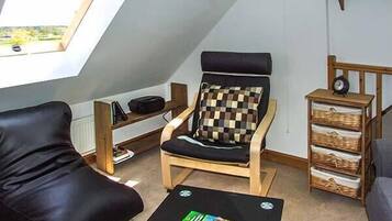 A cozy room with a black leather sofa, a wooden armchair with a checkered cushion, a small black table with magazines, a wooden shelf with a globe and books, and a skylight above.