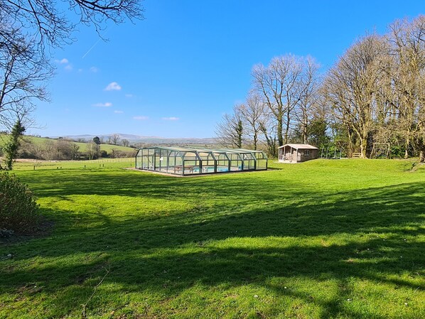 A greenhouse structure with a glass roof, surrounded by a grassy field and trees. - Swallow Cottage (Whitland)