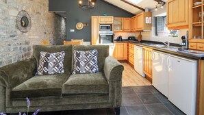 A cozy living room with a two-seater sofa and a stone wall. A kitchen with wooden cabinets and a dishwasher is visible. - Pear Tree Cottage (Wedmore)