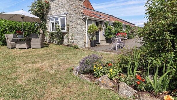 A stone house with a tiled roof, a patio with outdoor furniture, and a garden with various plants and flowers. - Pear Tree Cottage (Wedmore)