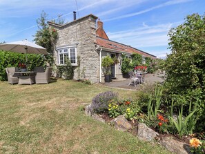 A stone house with a tiled roof, a patio with outdoor furniture, and a garden with various plants and flowers. - Pear Tree Cottage (Wedmore)