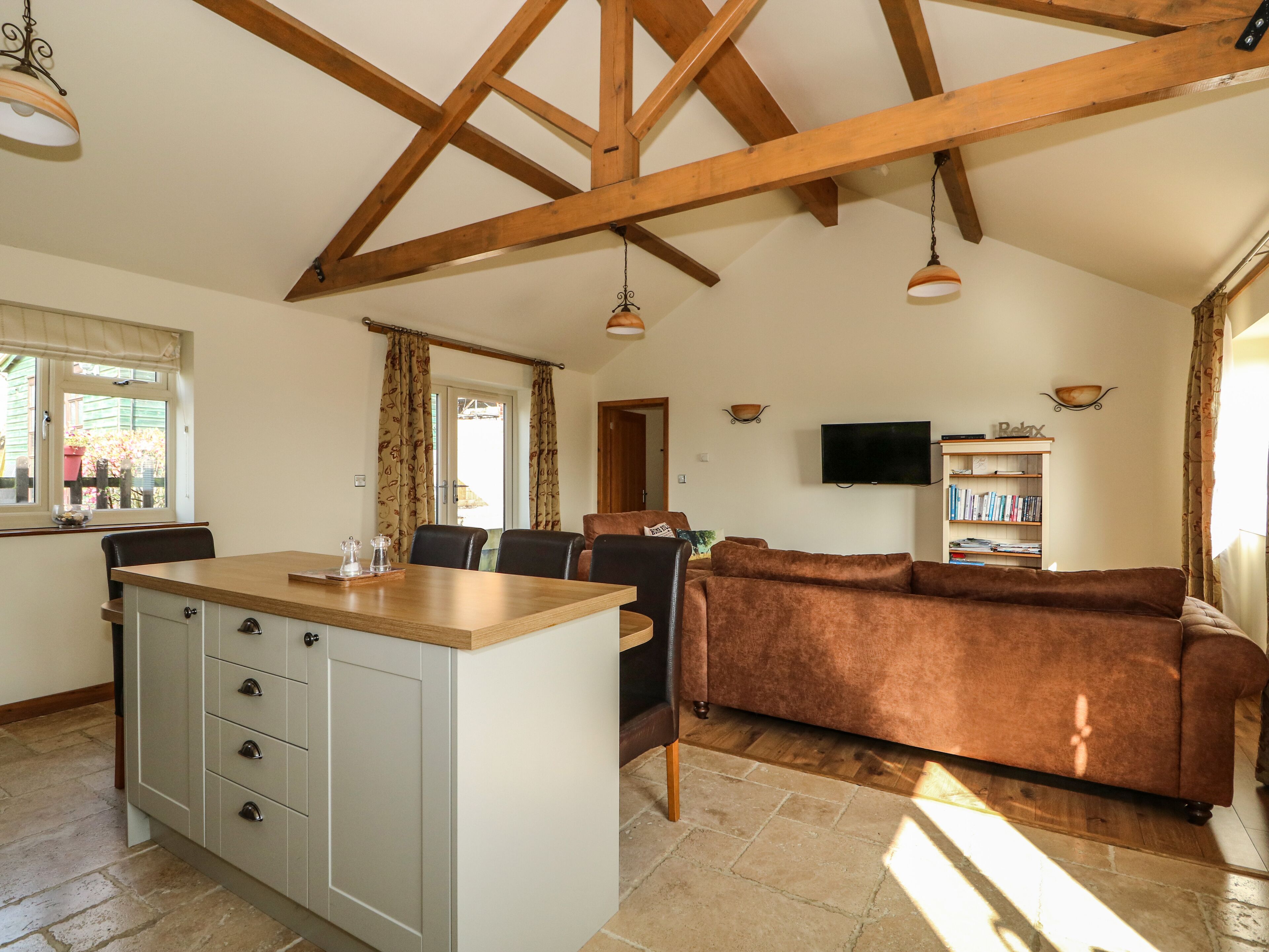 A spacious living area with a brown sofa, wooden ceiling beams, a kitchen island with cabinets, a dining table, and a TV mounted on the wall.