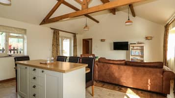 A spacious living area with a brown sofa, wooden ceiling beams, a kitchen island with cabinets, a dining table, and a TV mounted on the wall.