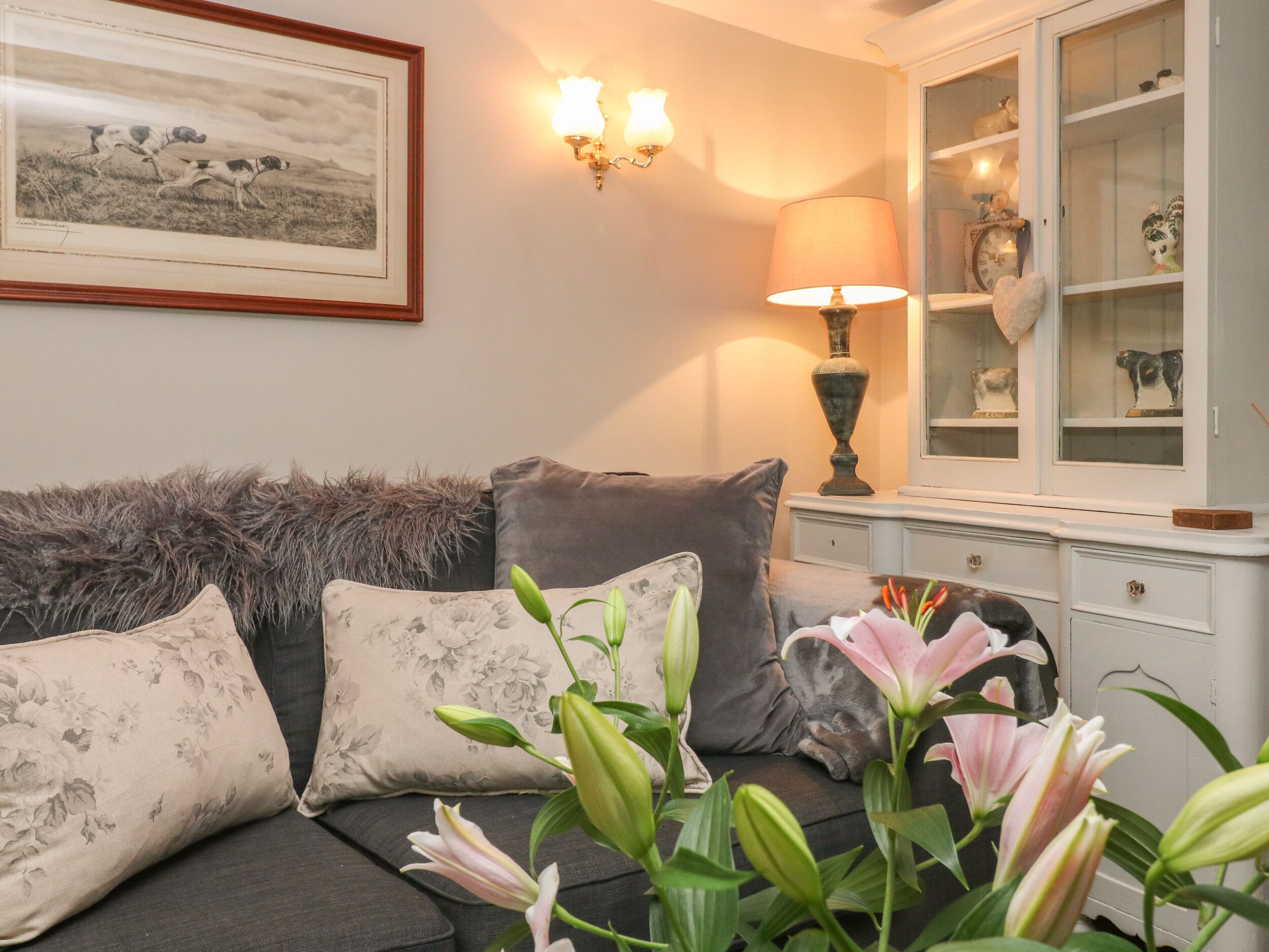 A living room with a sofa, floral cushions, a lamp, a cabinet with glass doors, and a framed picture on the wall.