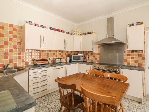 A kitchen with white cabinets, a wooden table, and a tiled backsplash. - Minffordd (Llanfairpwllgwyngyll)