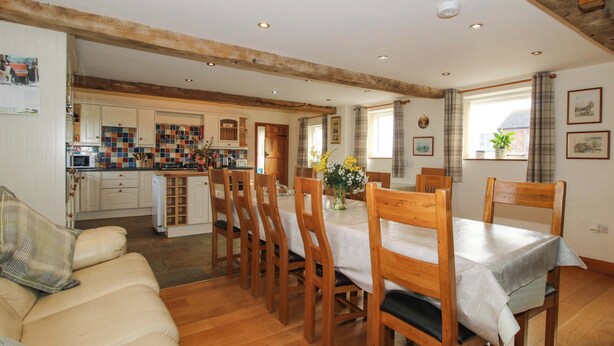 A cozy living room with a sofa, wooden dining table, and chairs, a kitchen with a tiled backsplash, and a wooden ceiling beam. - The Barn (Ellesmere)
