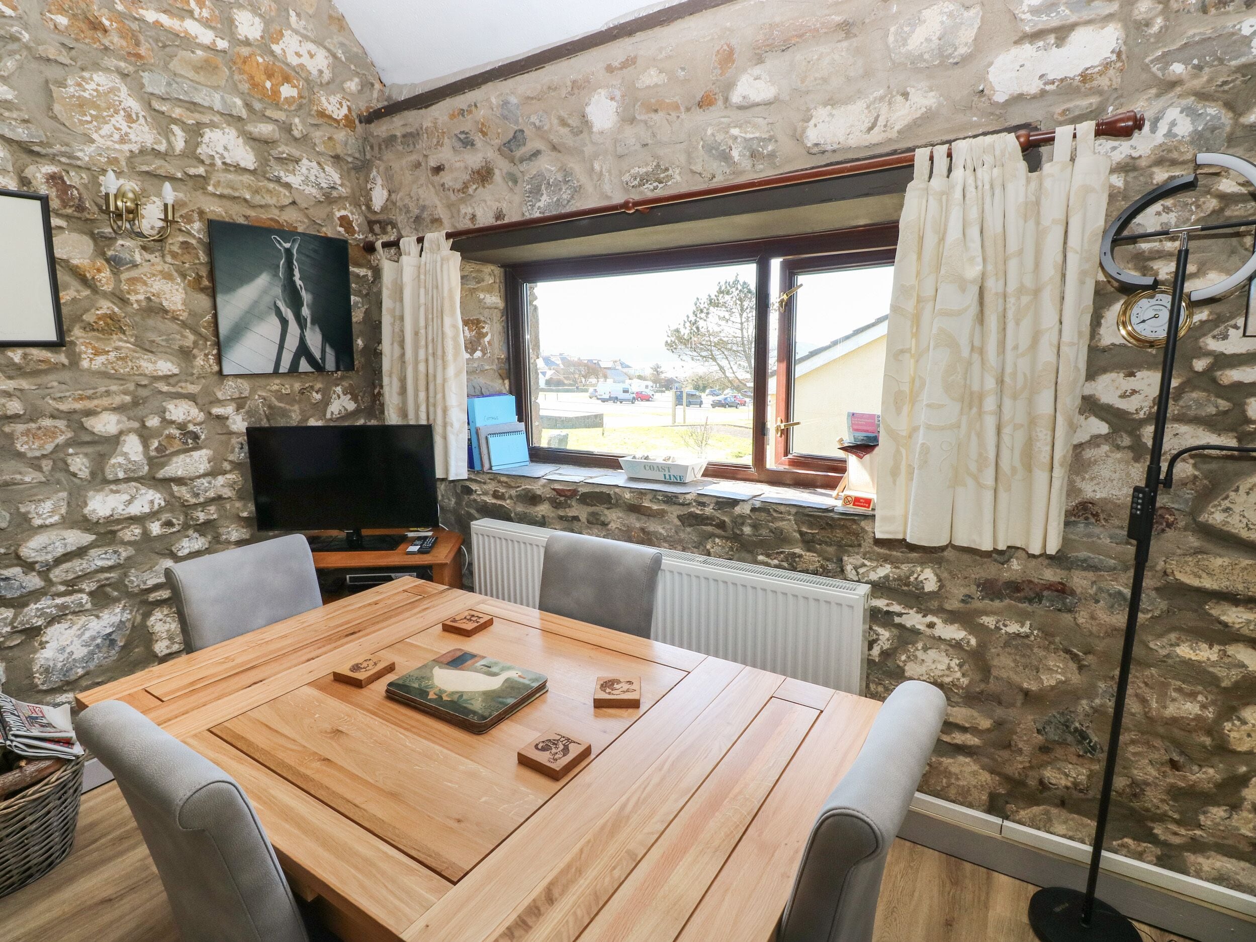 A stone-walled room with a wooden dining table, chairs, a television, and a window with curtains.