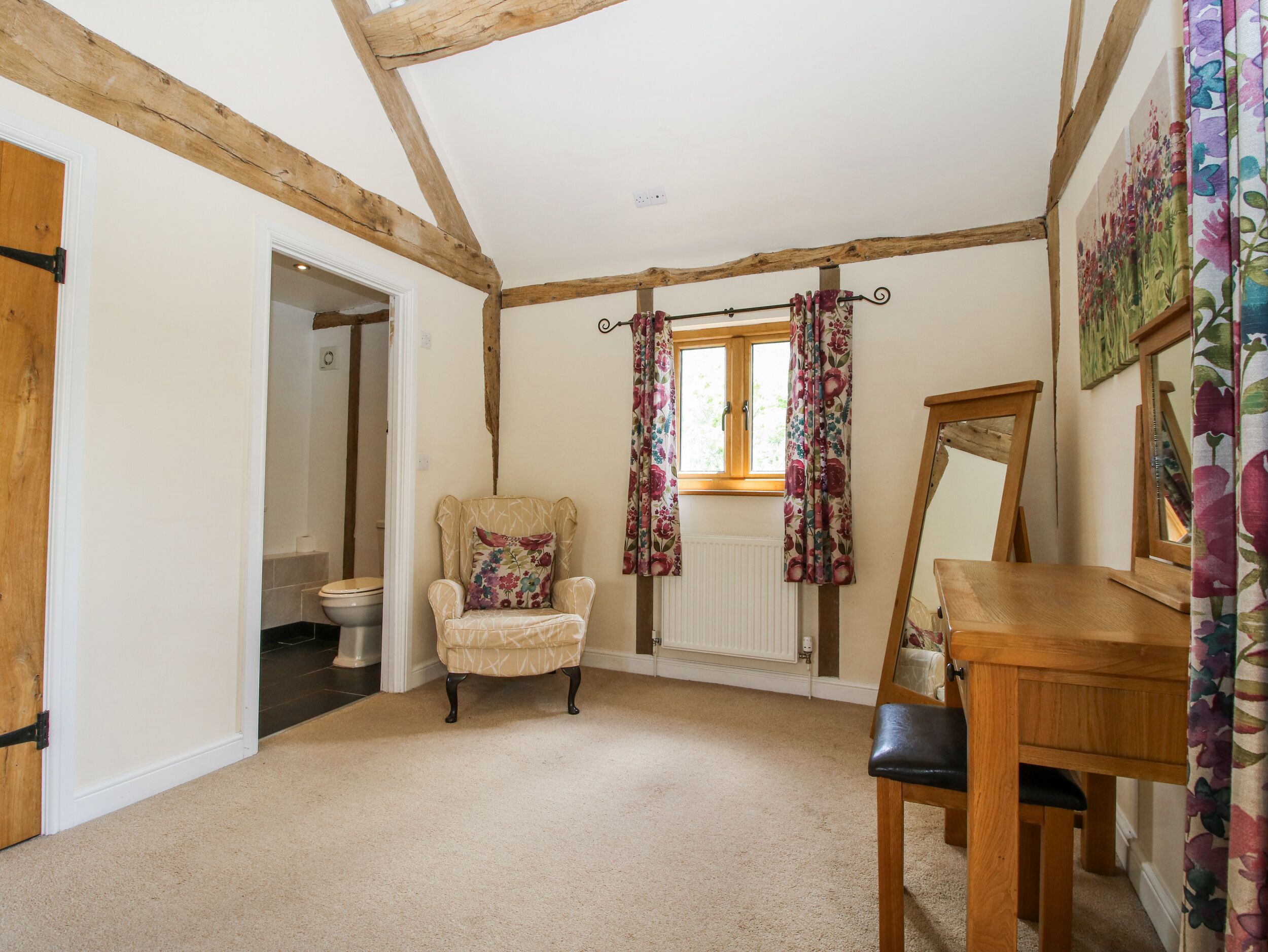 A room with exposed wooden beams, a wooden chair with a floral cushion, a wooden table, and a window with floral curtains.