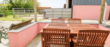 Um pátio externo com mesa e cadeiras de madeira, um banco de metal e uma parede rosa.
