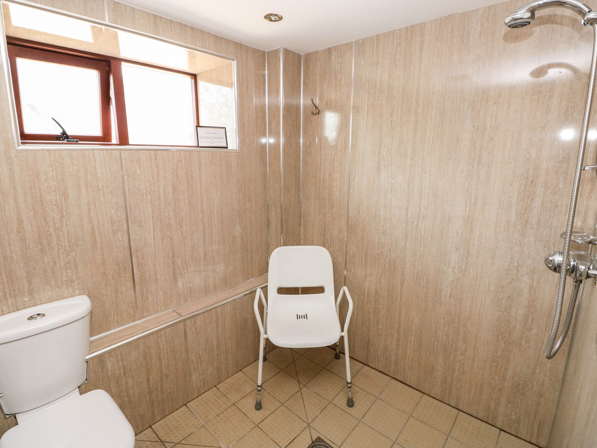 A small, tiled bathroom with a toilet, a shower, and a white chair.