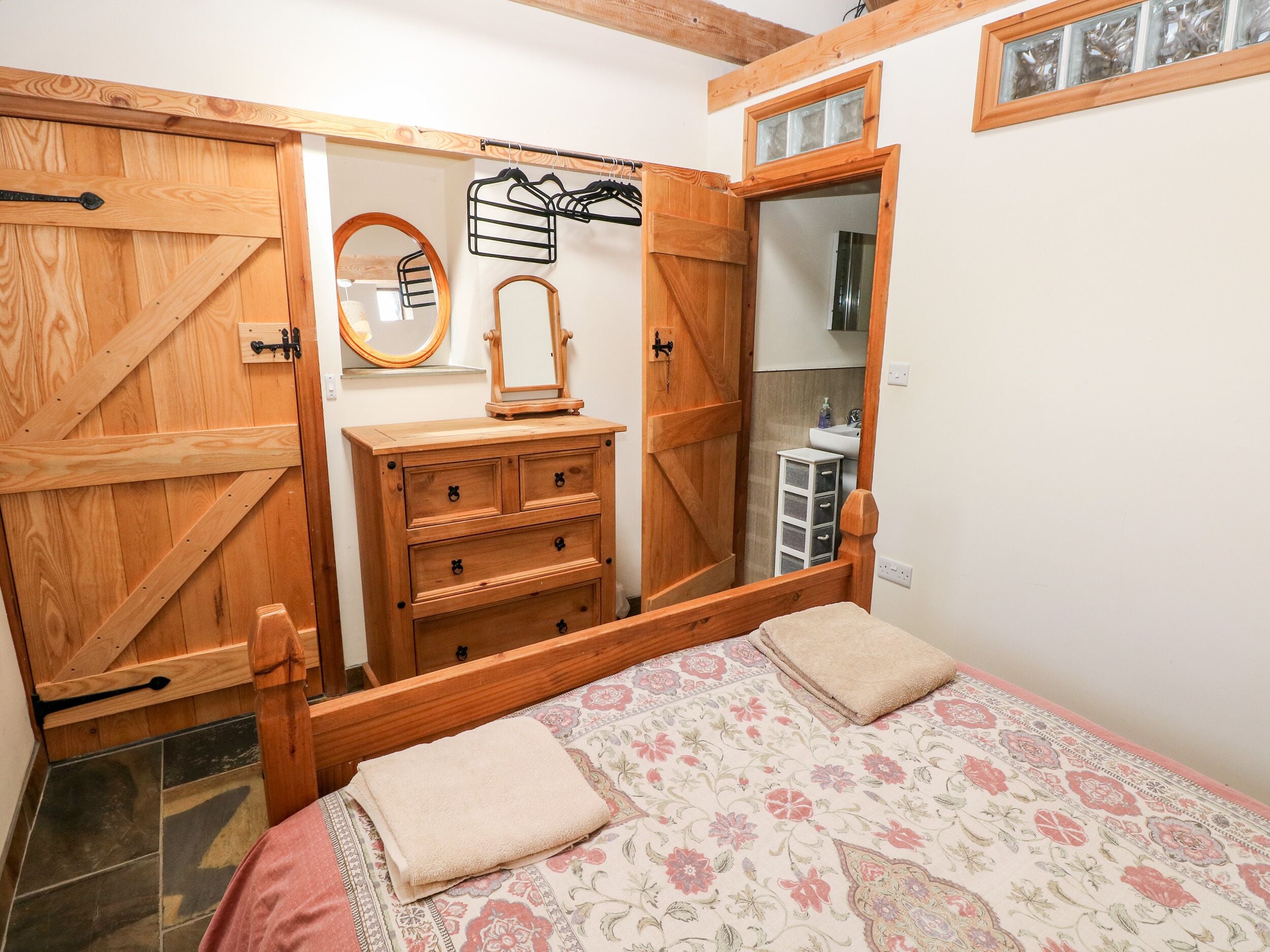 A bedroom with a wooden wardrobe, a dresser, a mirror, and a bed with floral bedding.