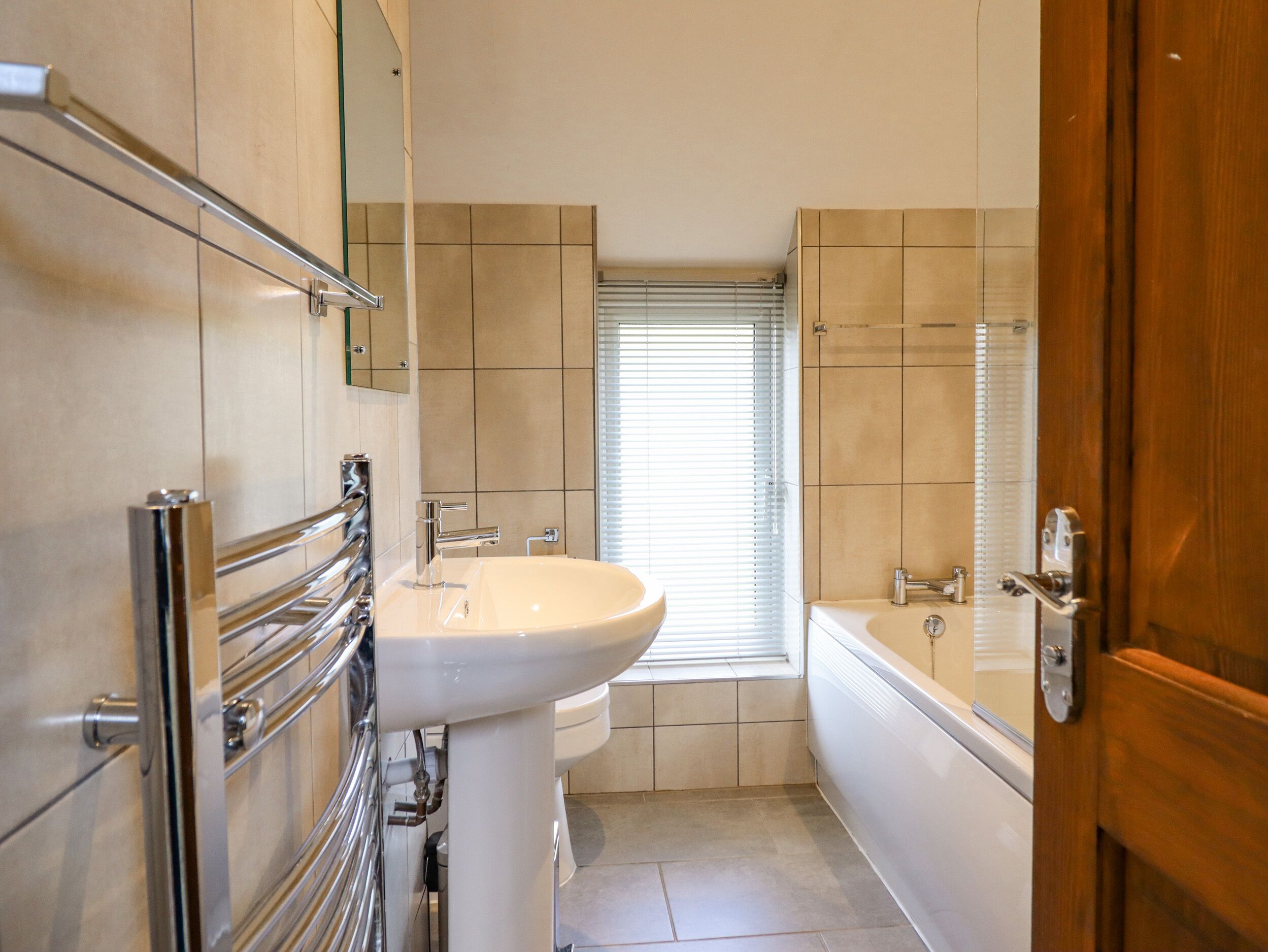 Une salle de bain avec un lavabo blanc, des robinetteries chromées, une fenêtre avec des stores et une porte en bois.