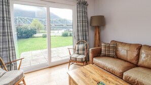 Sala de estar com sofá, mesa de centro de madeira, cadeira e luminária. Há uma janela grande com cortinas, e vista para uma ponte e vegetação lá fora.
