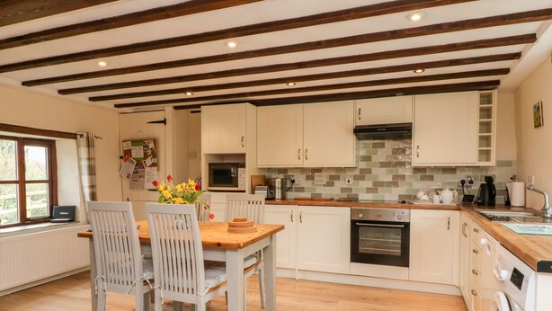 A modern kitchen with wooden beams, white cabinets, and a wooden dining table with chairs. - Owl's Nest (South Molton)