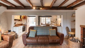 A cozy living room with a brown leather sofa, a wooden ceiling, and a kitchen area visible through an open doorway. - Woodbine Cottage (Market Rasen)