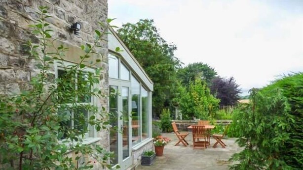 A stone building with a glass door and windows, a concrete patio, and a wooden table and chairs. - The Granary (York)