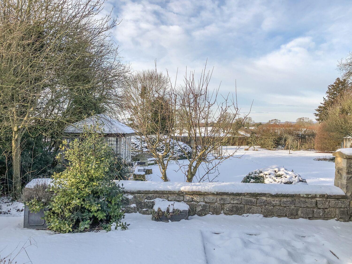 Un jardín cubierto de nieve con un muro de piedra, un pabellón y árboles sin hojas.