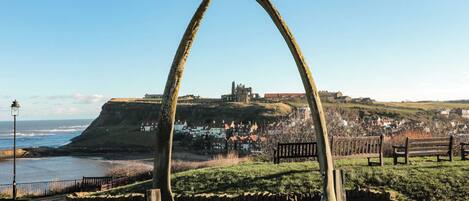 A historic stone archway with a bird perched on top, overlooking a coastal town with a prominent castle in the background.