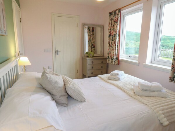 A bedroom with a bed, a dresser, a mirror, and a window with curtains. - Driftwood Cottage (Isle of Mull)