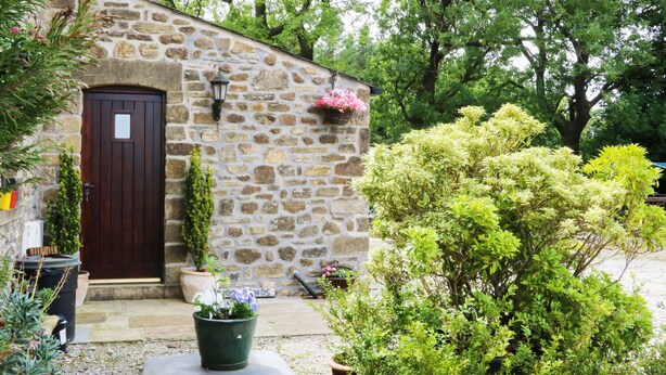 A stone building with a dark wooden door, a small porch, and potted plants. - Woodlands (Skipton)