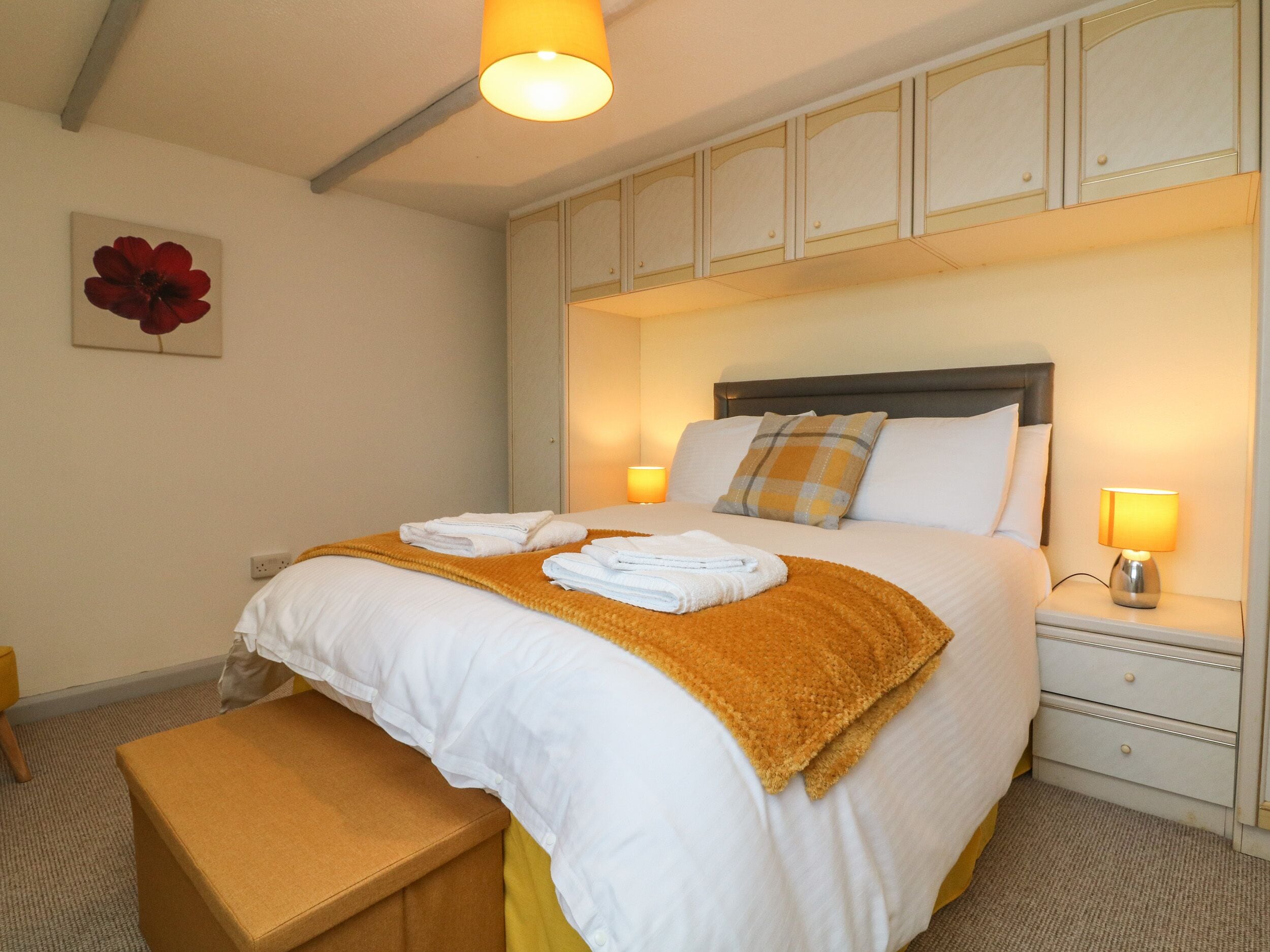 A neatly made bed with white and yellow bedding, a bench, and a bedside table with a lamp.