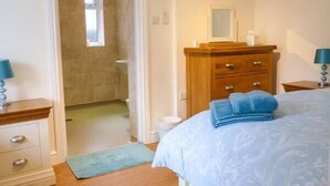 A bedroom with a bed, a wooden dresser, a nightstand, a bathroom visible through an open door, and a blue rug on the floor. - The Old Dairy (Beverley)