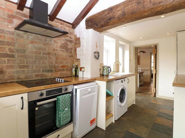 A compact kitchen with a brick wall, wooden beams, a built-in oven, a dishwasher, and a washing machine. - Fern Cottage (King's Lynn)