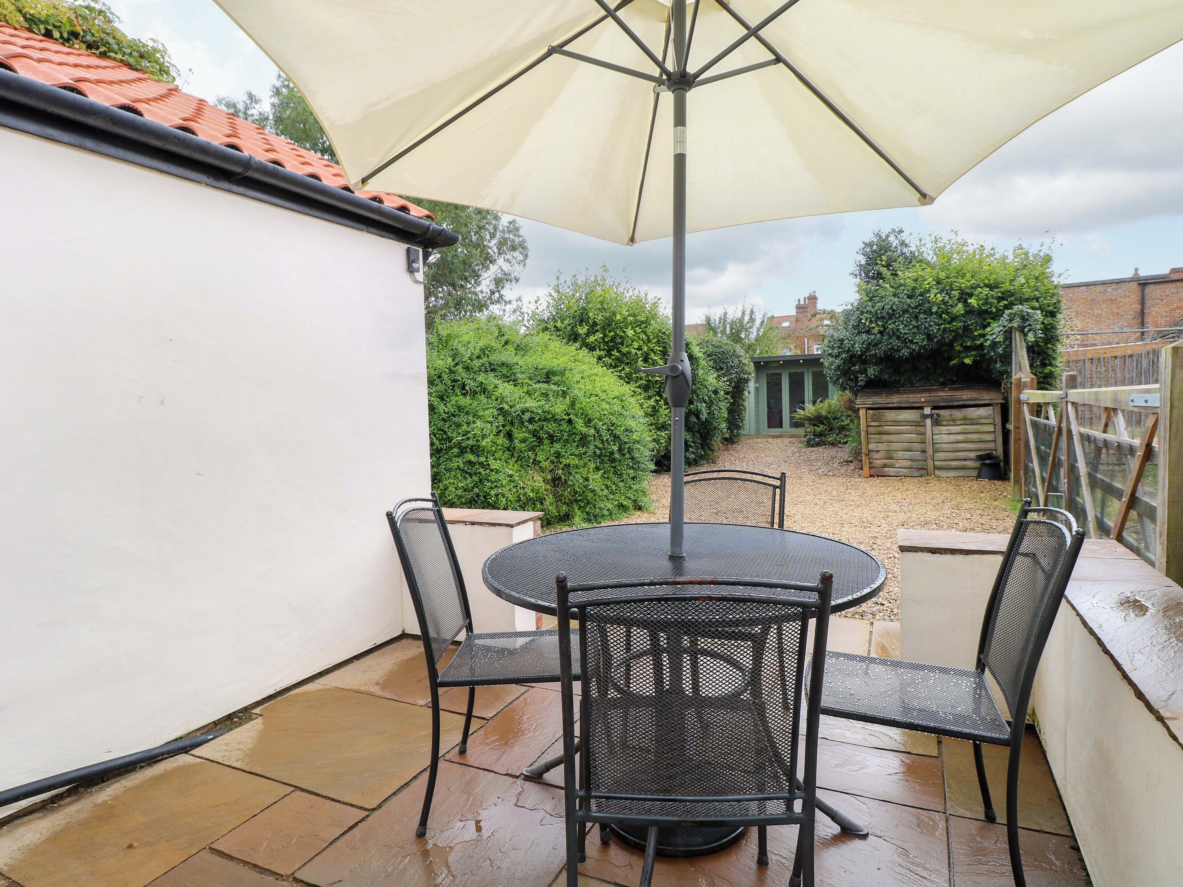 An outdoor patio with a round table and four chairs, a large umbrella, and a view of a garden and neighboring buildings.