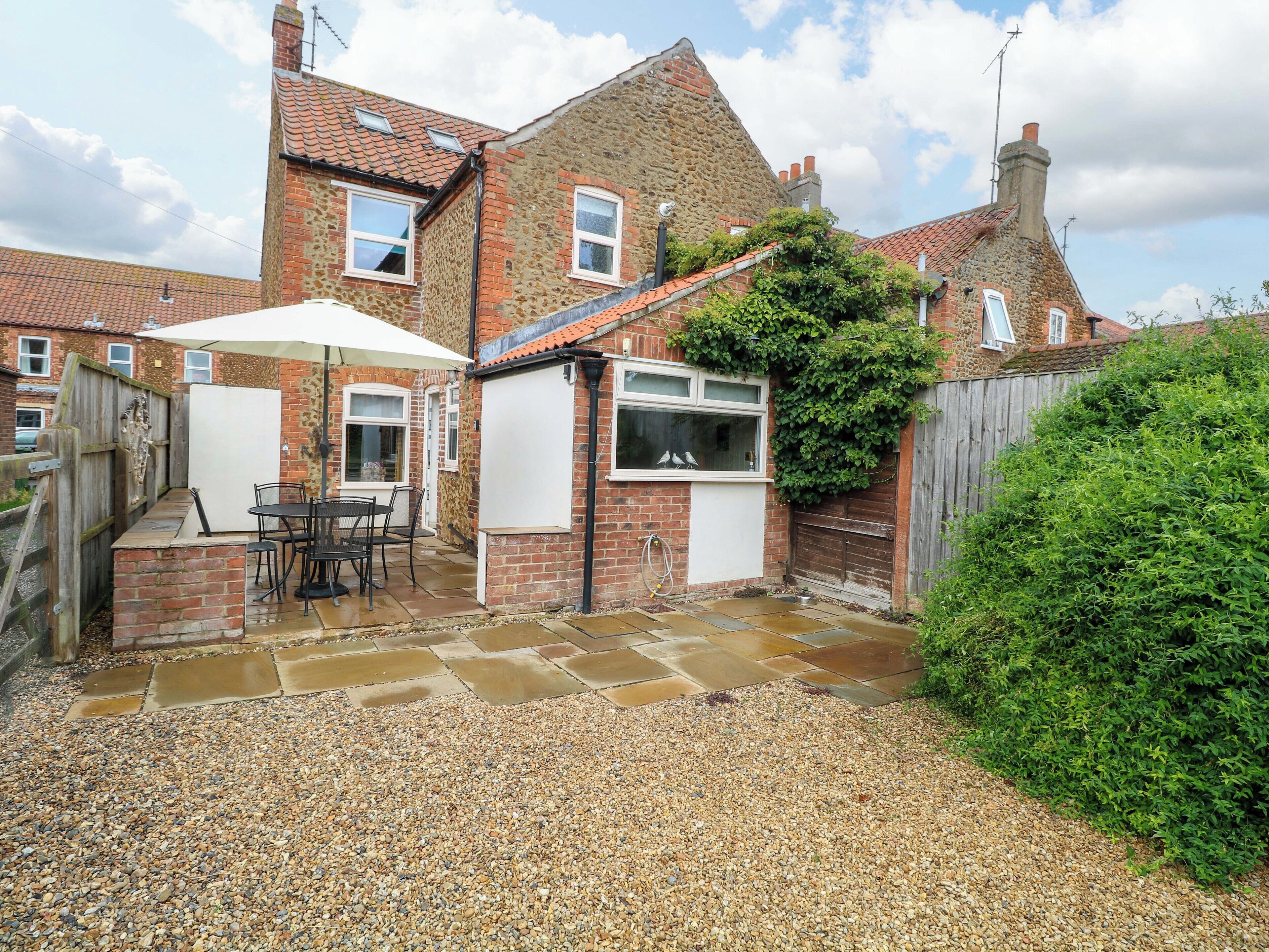 A two-story house with a patio area featuring a table and chairs, a small enclosed space with a window, and a garden with a wooden fence.