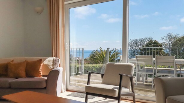 A living room with a sofa, a table, and a chair, a balcony with a view of the sea, and outdoor furniture. - Treverrion (St Austell)